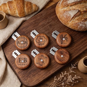 Puede incluir: Colección de cuchillas para pan de madera con diseños y nombres grabados. Las cuchillas tienen una hoja plateada unida a un mango redondo de madera. Los mangos están grabados con texto e imágenes, incluyendo un gorro de chef, trigo e iniciales.