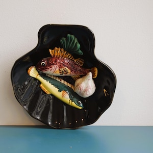 Peut inclure: Plat en céramique noir en forme de coquillage avec un motif décoratif. Le plat présente un poisson rouge et jaune, un poisson vert et jaune, une gousse d'ail et des accents d'algues vertes.
