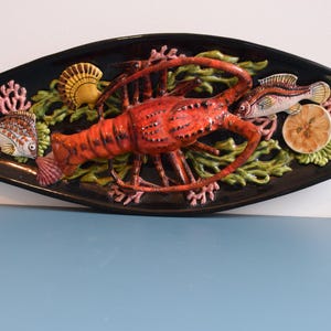 Puede incluir: Una fuente ovalada de cerámica negra con un diseño en relieve que presenta una langosta roja, peces, una concha y coral. La langosta está en el centro, rodeada de algas verdes y otras criaturas marinas. La fuente es decorativa.
