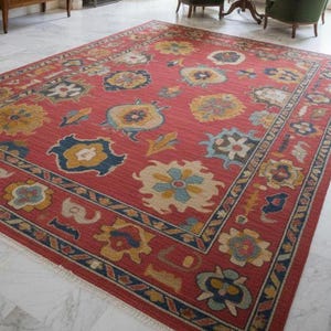 Könnte beinhalten: Ein großer, roter Teppich mit floralem Muster dominiert den Raum. Der Teppich hat eine Bordüre und sich wiederholende Blumenmotive in Blau, Gelb und Creme. Der Raum umfasst auch antike Möbel, Gemälde und einen Kronleuchter.