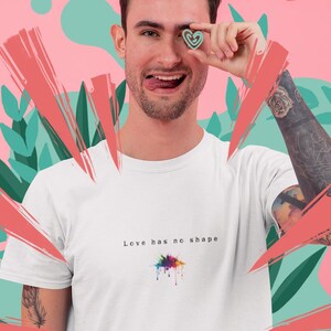 May include: A white short-sleeved t-shirt featuring the black text "Love has no shape" printed on the chest above a small, colorful, multi-hued paint splatter graphic. The shirt is made of a plain fabric and has a standard crew neckline.