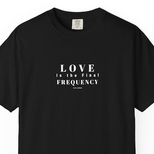May include: A black short-sleeve t-shirt featuring white text centered on the chest that reads LOVE IS THE FINAL FREQUENCY EST.2024. The shirt has a crew neckline and a standard fit, with a small white brand label visible inside the collar area.