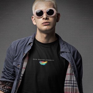 May include: A black t-shirt featuring the text Love beyond judgment and a small rainbow graphic, worn with a blue button-down shirt with a plaid lining, light blue denim jeans, a brown belt, and black boots, paired with round pink-tinted sunglasses.
