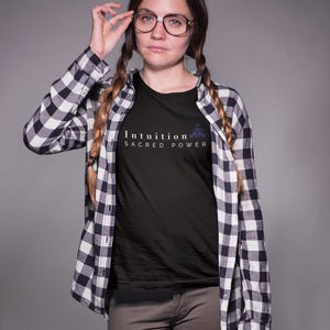 May include: Black t-shirt with the words "Intuition Sacred Power" in white. The shirt is worn under a black and white plaid button-down shirt. The person is wearing glasses and brown pants.