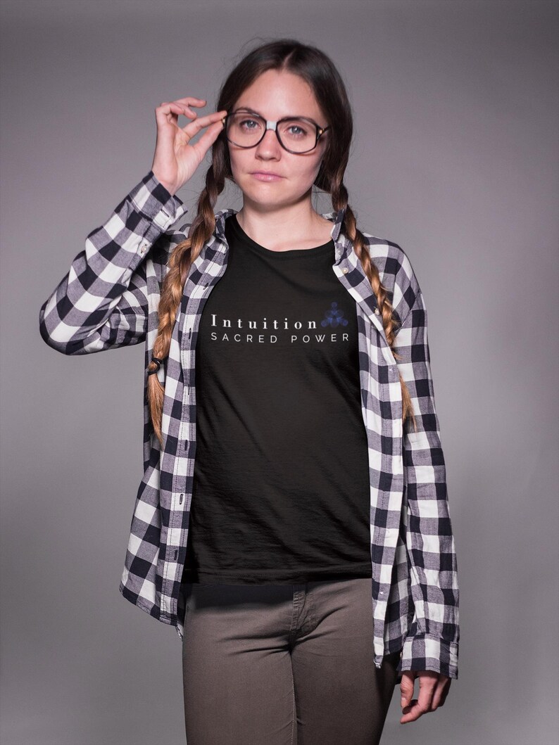 May include: Black t-shirt with the words "Intuition Sacred Power" in white. The shirt is worn under a black and white plaid button-down shirt. The person is wearing glasses and brown pants.