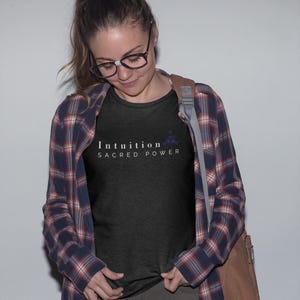 May include: A black t-shirt with the words "Intuition Sacred Power" in white text and a small blue floral design. The person is wearing a plaid shirt over the t-shirt, glasses, and a brown shoulder bag.