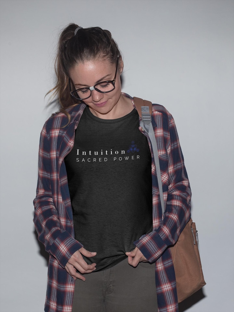 May include: A black t-shirt with the words "Intuition Sacred Power" in white text and a small blue floral design. The person is wearing a plaid shirt over the t-shirt, glasses, and a brown shoulder bag.