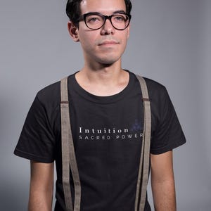 May include: A person wearing black-framed glasses, a black t-shirt with the text "Intuition Sacred Power", and suspenders. The suspenders are a light brown color with a herringbone pattern. The background is a solid gray.