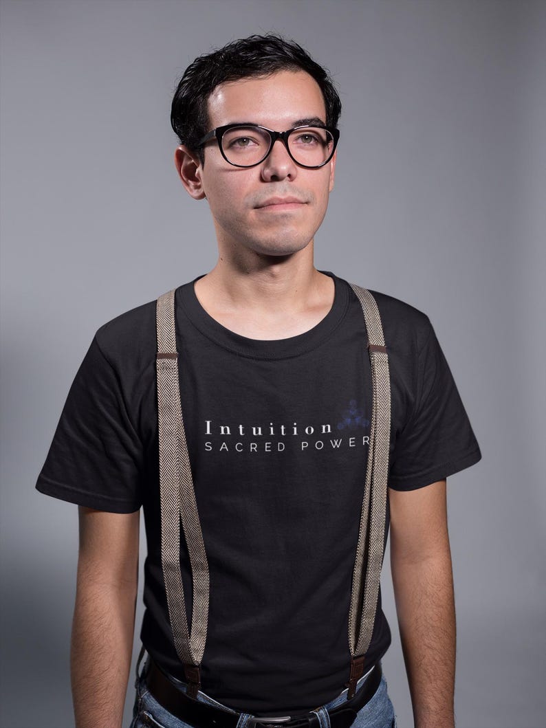 May include: A person wearing black-framed glasses, a black t-shirt with the text "Intuition Sacred Power", and suspenders. The suspenders are a light brown color with a herringbone pattern. The background is a solid gray.