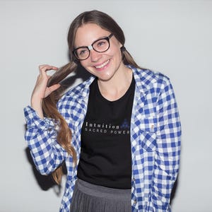 May include: A woman wearing a black t-shirt with the text "Intuition Sacred Power" in white, paired with a blue and white checkered shirt. She is also wearing glasses and smiling.
