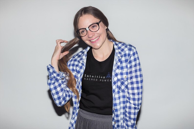 May include: A woman wearing a black t-shirt with the text "Intuition Sacred Power" in white, paired with a blue and white checkered shirt. She is also wearing glasses and smiling.