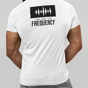 Puede incluir: Camiseta blanca con un gráfico rectangular negro en la espalda. El gráfico presenta un diseño de onda de sonido sobre las palabras "YOU ARE THE FREQUENCY" en negrita, con fuente sans-serif. La camiseta es de manga corta y parece estar hecha de un material suave.