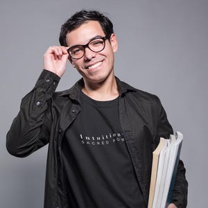 May include: A person wearing a black shirt and jacket, holding books and glasses. The black t-shirt has the words "Intuition Sacred Power" printed on it in white. The person is smiling and looking at the camera.