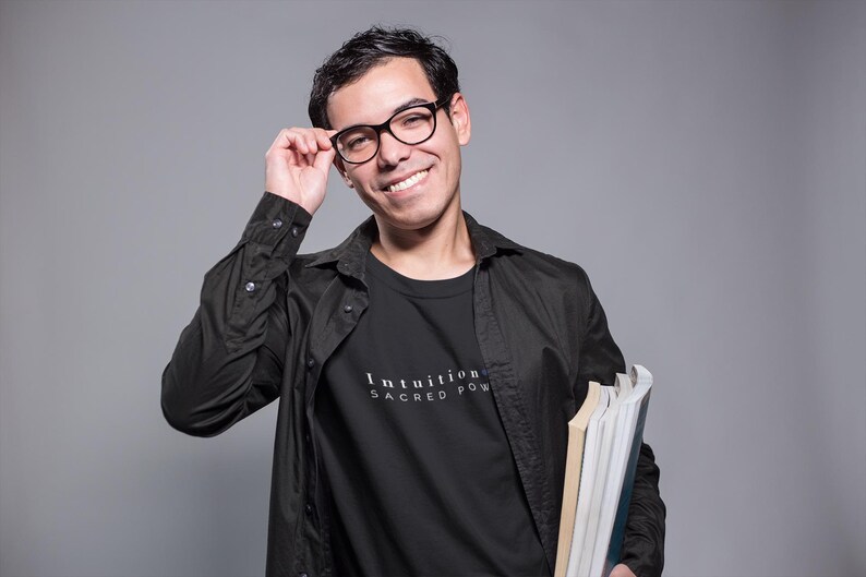 May include: A person wearing a black shirt and jacket, holding books and glasses. The black t-shirt has the words "Intuition Sacred Power" printed on it in white. The person is smiling and looking at the camera.