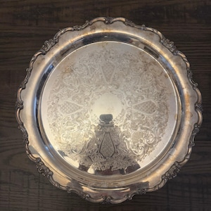May include: A round, silver-colored serving tray with an ornate, embossed floral design. The tray has a scalloped edge and a reflective surface, suitable for serving food or displaying decorative items.