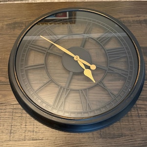 May include: A round, black wall clock with Roman numerals and gold-colored hands. The clock face is visible through a clear glass cover. The clock is a decorative item for home decor.