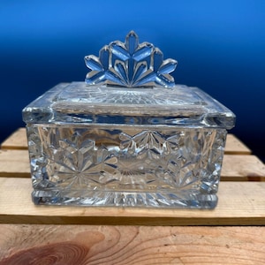 May include: A clear, square glass trinket box with a decorative lid and a floral-shaped handle. The box features a cut-glass design with starburst and geometric patterns. The box is displayed on a wooden surface against a blue backdrop.
