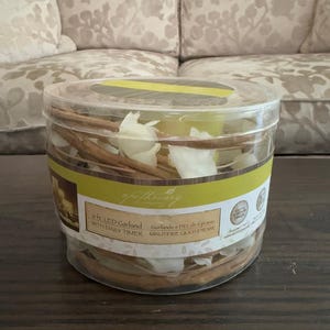 May include: A clear plastic container holding a 6 ft. LED garland with a daily timer. The garland includes brown sticks and white floral accents. The container has a yellow band and the word "apothecary" is visible.