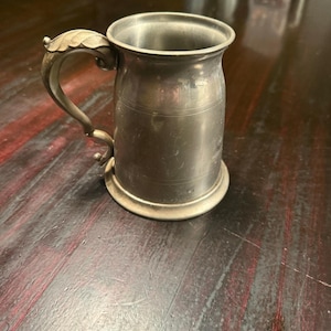 May include: A vintage pewter mug with a decorative handle. The mug has a cylindrical body with a flared base and a detailed handle. The handle features a leaf-like design. The mug is made of a silver-colored metal and appears to be well-worn.