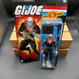 May include: A G.I. Joe Destro action figure in a plastic package. The package features the G.I. Joe logo, the word "Destro", and a graphic of the character. The figure is wearing a black outfit with a red collar and silver accents. Includes a briefcase and weapon.