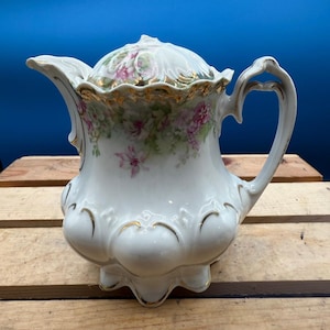 May include: A white porcelain creamer with a lid, featuring a floral design in pink and green. The creamer has a gold trim and a decorative handle. The creamer is sitting on a wooden surface.