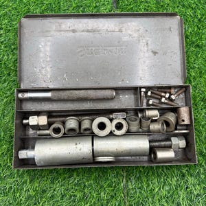 May include: An open, gray metal toolbox filled with various metal tools. The tools include a handle, bolts, and several cylindrical and circular pieces. The toolbox is sitting on a bed of green grass.