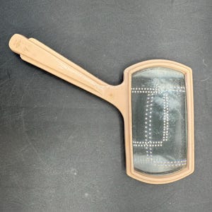 May include: A vintage, peach-colored hand mirror with a rectangular mirror surface. The handle is attached to a frame that surrounds the mirror. The mirror surface has a pattern of small white dots. The mirror is slightly dusty.