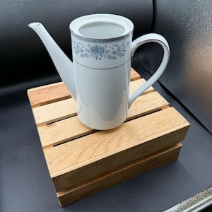 May include: White porcelain coffee server with a long spout and handle. The server has a floral blue and silver design around the rim. It is sitting on a small wooden crate.