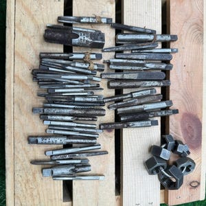 May include: A collection of metal taps and dies, used for cutting threads, arranged on a wooden pallet. The tools are various sizes and show signs of use. Several hex nuts are also present.