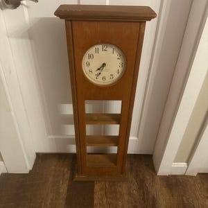 May include: A tall, wooden clock with a round clock face and Roman numerals. The clock is brown and has a rectangular body with three shelves. The clock is standing on a wooden floor.