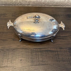 May include: A silver-colored, oval serving dish with a lid, ornate handles, and a decorative knob. The dish is elevated on small feet and reflects the surrounding environment. The dish is likely vintage and suitable for serving food.