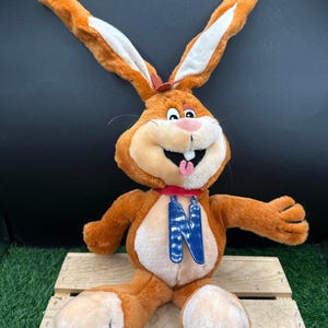 May include: A plush brown rabbit with long ears, a pink nose, and a red collar. The rabbit has a blue pendant with the letter "N" on it. It is sitting on a wooden pallet.