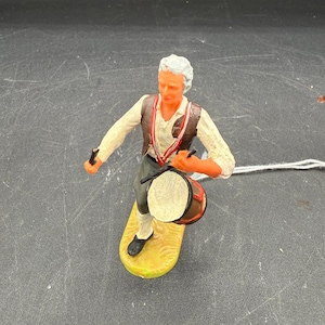 May include: A small figurine of a drummer in a white shirt, brown vest, and black pants. The drummer is playing a small drum with a red rim. The figurine is mounted on a yellow base.