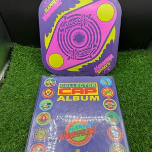May include: A purple and pink Slammer Whammers Face-Off game arena and a Collector Cap Album. The game arena is a square shape with a circular design and the words "Face-Off" and "Slammer Whammers". The album is purple with colorful cap designs.