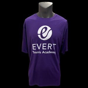 Puede incluir: Una camiseta morada con el texto blanco "EVERT Tennis Academy" y un logotipo blanco. El logotipo es un círculo blanco con una "e" blanca estilizada en su interior. La camiseta se muestra en un maniquí sobre un fondo negro.