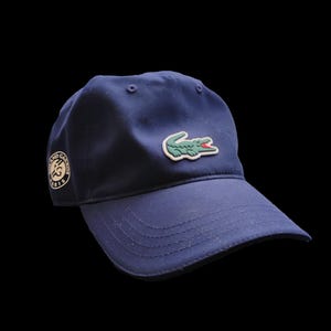 Könnte beinhalten: Marineblaue Baseballkappe mit dem ikonischen grünen Krokodil-Logo auf der Vorderseite. Die Kappe hat auch einen runden Aufnäher mit Text an der Seite. Ein klassisches, sportliches Accessoire.