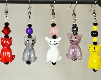 Può includere: Cinque paia di orecchini di vetro a forma di gatto con perline di diversi colori. Gli orecchini sono rossi, grigi, bianchi, viola e gialli.