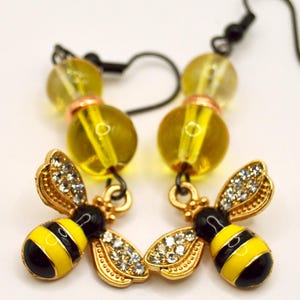 Pu&ograve; includere: Un paio di orecchini a tema ape. Ogni orecchino presenta un ciondolo a forma di ape color oro con strisce nere e gialle e accenti simili a cristalli sulle ali. Gli orecchini hanno anche accenti di perline gialle e ganci neri.