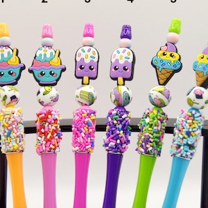 May include: A collection of six colorful pens with decorative toppers. Each pen features a different design, including cupcakes, ice cream cones, and popsicles. The pen barrels are covered in colorful sprinkles, and the toppers have a variety of colors.