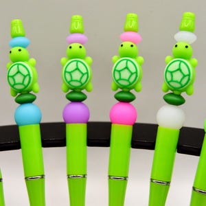 May include: Six green pens with turtle-shaped toppers. Each pen has a different coloured accent, including yellow, blue, purple, pink, white, and green. The turtle toppers feature a green shell design.