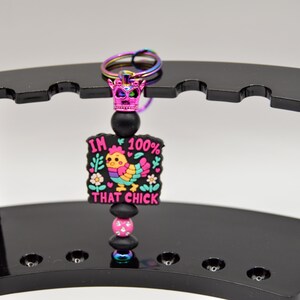 May include: A black and pink keychain with a colorful cartoon chicken and the text "I'm 100% That Chick". The keychain has a black and pink beaded charm and a rainbow crown.