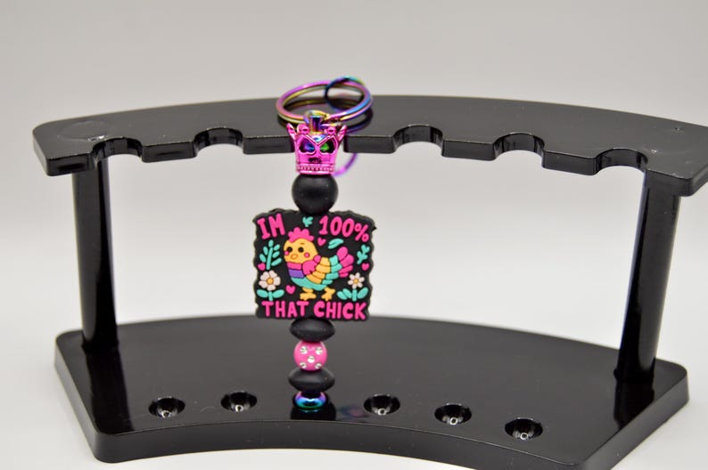 May include: A black and pink keychain with a colorful cartoon chicken and the text "I'm 100% That Chick". The keychain has a black and pink beaded charm and a rainbow crown.