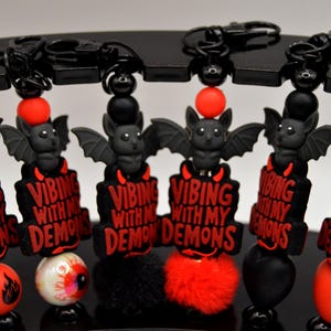 May include: Black keychains with a bat and the text "VIBING WITH MY DEMONS" in red. Each keychain has a different charm, including a red ball, an eyeball, a black pom-pom, a red pom-pom, and a black heart.
