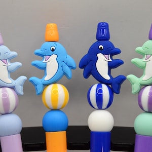 May include: Four colorful dolphin-themed pen toppers. Each dolphin is a different color: light blue, blue, dark blue, and green. Each dolphin has a small bucket on its head. The pens have striped and solid colored beads.