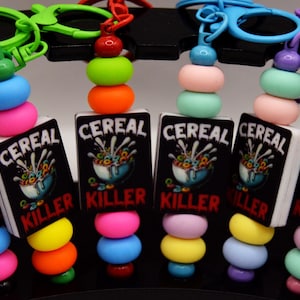 May include: A collection of colorful keychains featuring a small, rectangular design with the words "CEREAL KILLER" on a black background. Each keychain has a stack of colorful beads and a matching clasp.