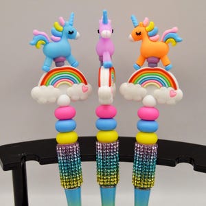 Könnte beinhalten: Drei Stifte mit Einhorn-Aufsätzen. Jeder Stift hat ein andersfarbiges Einhorn auf einem Regenbogen- und Wolken-Design. Die Stiftkörper sind mit farbigen Strasssteinen verziert. Die Stifte werden auf einem schwarzen Ständer ausgestellt.