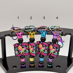 May include: Four colorful keychains with a black and pink charm featuring a cartoon chicken and the text "I'm 100% That Chick". Each keychain has a rainbow iridescent metal ring and a black bead with a crown charm.