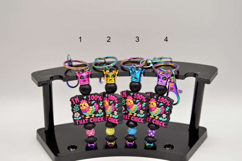 May include: Four colorful keychains with a black and pink charm featuring a cartoon chicken and the text "I'm 100% That Chick". Each keychain has a rainbow iridescent metal ring and a black bead with a crown charm.