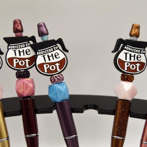 May include: A collection of pens with decorative tops. Each pen features a coffee pot design with the text "Addicted to The Pot". The pens have various colors, including gold, brown, and pink, and are displayed on a black stand.