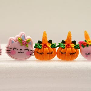 Puede incluir: Una colección de accesorios caprichosos con temática de Halloween. El surtido incluye caras de gato rosa con cuernos de unicornio, caras de calabaza naranja con los ojos cerrados y caras de unicornio rosa. Cada pieza está adornada con detalles florales y detalles.